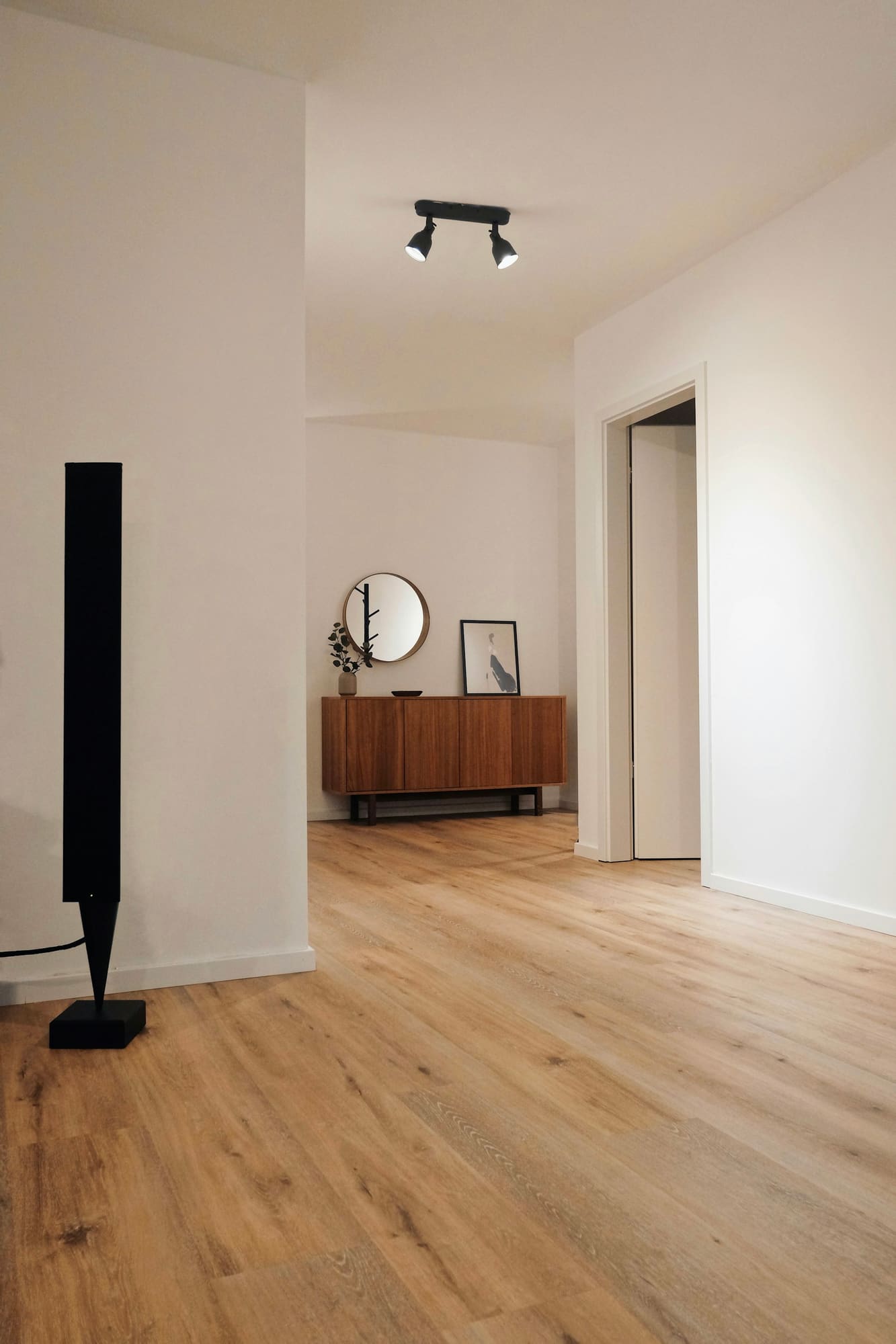Modern hallway with wooden flooring, white walls, mid-century credenza, round mirror, and black track lighting